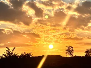 Por do sol. A imagem mostra o céu alaranjado, com algumas nuvens, com uma temperatura amena. Abaixo tem a sombra das arvores com um feixe de luz passando por elas.