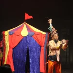 Pessoa se apresentando no Festival Internacional de Máscaras do Cariri. Na foto mostra um homem na frente de uma tenda colorida.