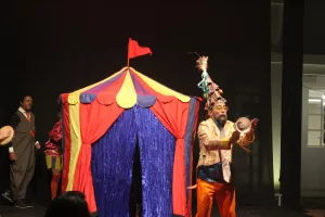 Pessoa se apresentando no Festival Internacional de Máscaras do Cariri. Na foto mostra um homem na frente de uma tenda colorida.