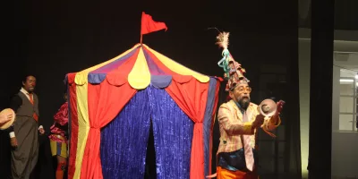 Pessoa se apresentando no Festival Internacional de Máscaras do Cariri. Na foto mostra um homem na frente de uma tenda colorida.
