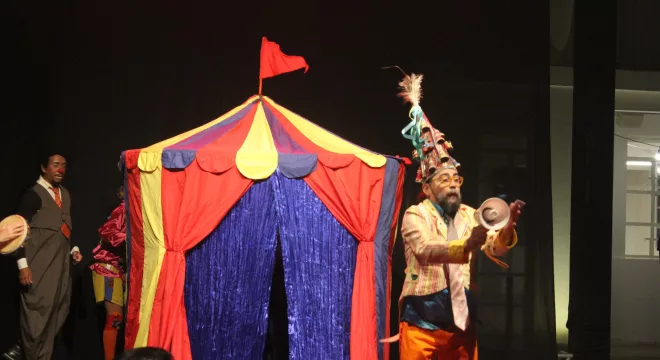 Pessoa se apresentando no Festival Internacional de Máscaras do Cariri. Na foto mostra um homem na frente de uma tenda colorida.