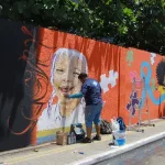 Parede sendo pintada com a técnica de grafite, em Juazeiro do Norte. A imagem mostra o artista próximo a parede pintando uma criança em um fundo laranja.