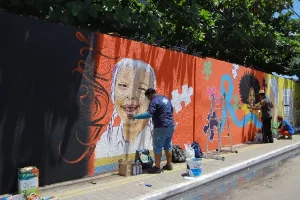 Parede sendo pintada com a técnica de grafite, em Juazeiro do Norte. A imagem mostra o artista próximo a parede pintando uma criança em um fundo laranja.