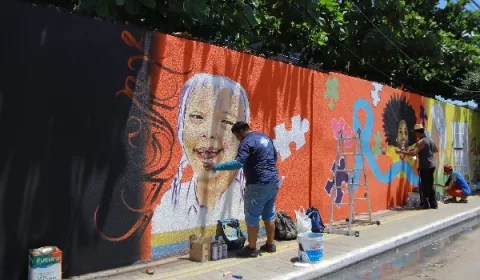 Parede sendo pintada com a técnica de grafite, em Juazeiro do Norte. A imagem mostra o artista próximo a parede pintando uma criança em um fundo laranja.