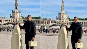 Padre segurando a mala bege com a Imagem Peregrina Mundial de Nossa Senhora de Fátima. Eçe está com uma roupa frente e em frente Santuário de Fátima, em Portugal.