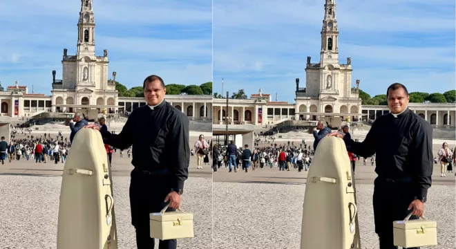 Padre segurando a mala bege com a Imagem Peregrina Mundial de Nossa Senhora de Fátima. Eçe está com uma roupa frente e em frente Santuário de Fátima, em Portugal.