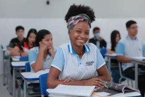 estudante juazeiro do norte sentada na cadeira em sala de aula. ela está sorrindo para a câmera e com os braços apoiados na mesa.