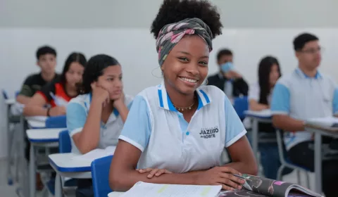 estudante juazeiro do norte sentada na cadeira em sala de aula. ela está sorrindo para a câmera e com os braços apoiados na mesa.