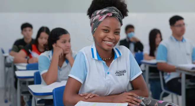 estudante juazeiro do norte sentada na cadeira em sala de aula. ela está sorrindo para a câmera e com os braços apoiados na mesa.