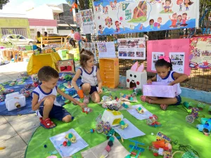 Crianças sentadas em um fundo verde com brinquedos sustentáveis desenvolvidos com material reciclado. Tem três crianças na imagem, durante a Brinquedoarte em Juazeiro do Norte.