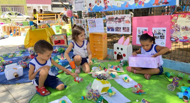 Crianças sentadas em um fundo verde com brinquedos sustentáveis desenvolvidos com material reciclado. Tem três crianças na imagem, durante a Brinquedoarte em Juazeiro do Norte.