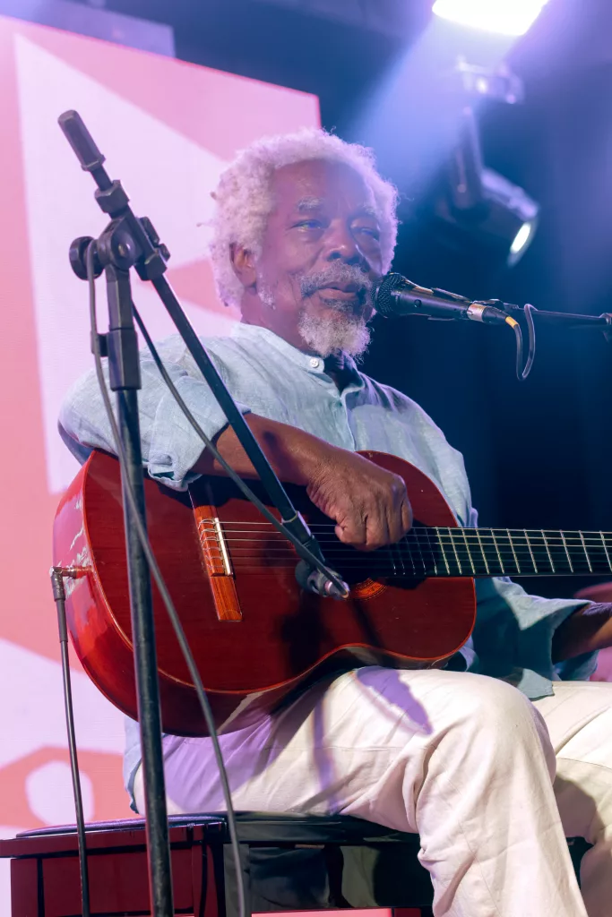 Mateus Aleluia se apresentando do Afrobarroco, no Cariri. Ele está sentado em uma cadeira, com o violão na mão.