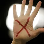 Mão com um x vermelho desenhado na palma, representando o pedido pelo fim da violência e feminicídio contra a mulher.