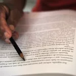 pessoa estudando. a imagem foca em uma página de livro onde a pessoa está segurando um lápis para acompanhar a leitura.