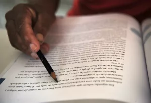 pessoa estudando. a imagem foca em uma página de livro onde a pessoa está segurando um lápis para acompanhar a leitura.