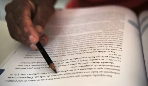 pessoa estudando. a imagem foca em uma página de livro onde a pessoa está segurando um lápis para acompanhar a leitura.