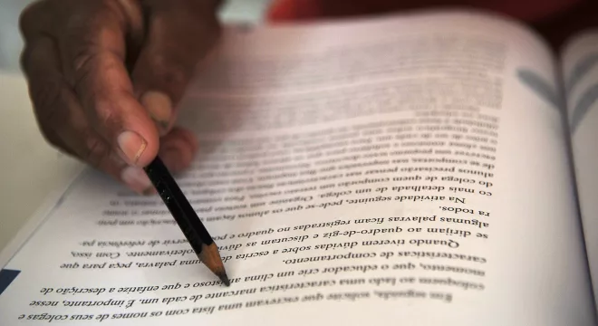 pessoa estudando. a imagem foca em uma página de livro onde a pessoa está segurando um lápis para acompanhar a leitura.